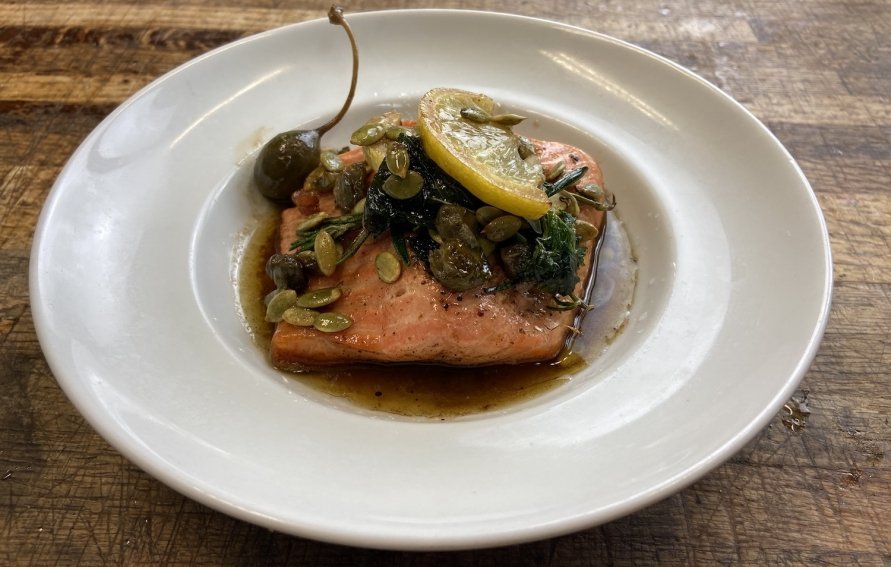 seared new york steelhead fish with lemon herbs and pepitas served on a white dinner plate