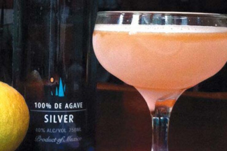 a peach colored cocktail in a coupe glass with a bottle of tequila and a lemon in the background